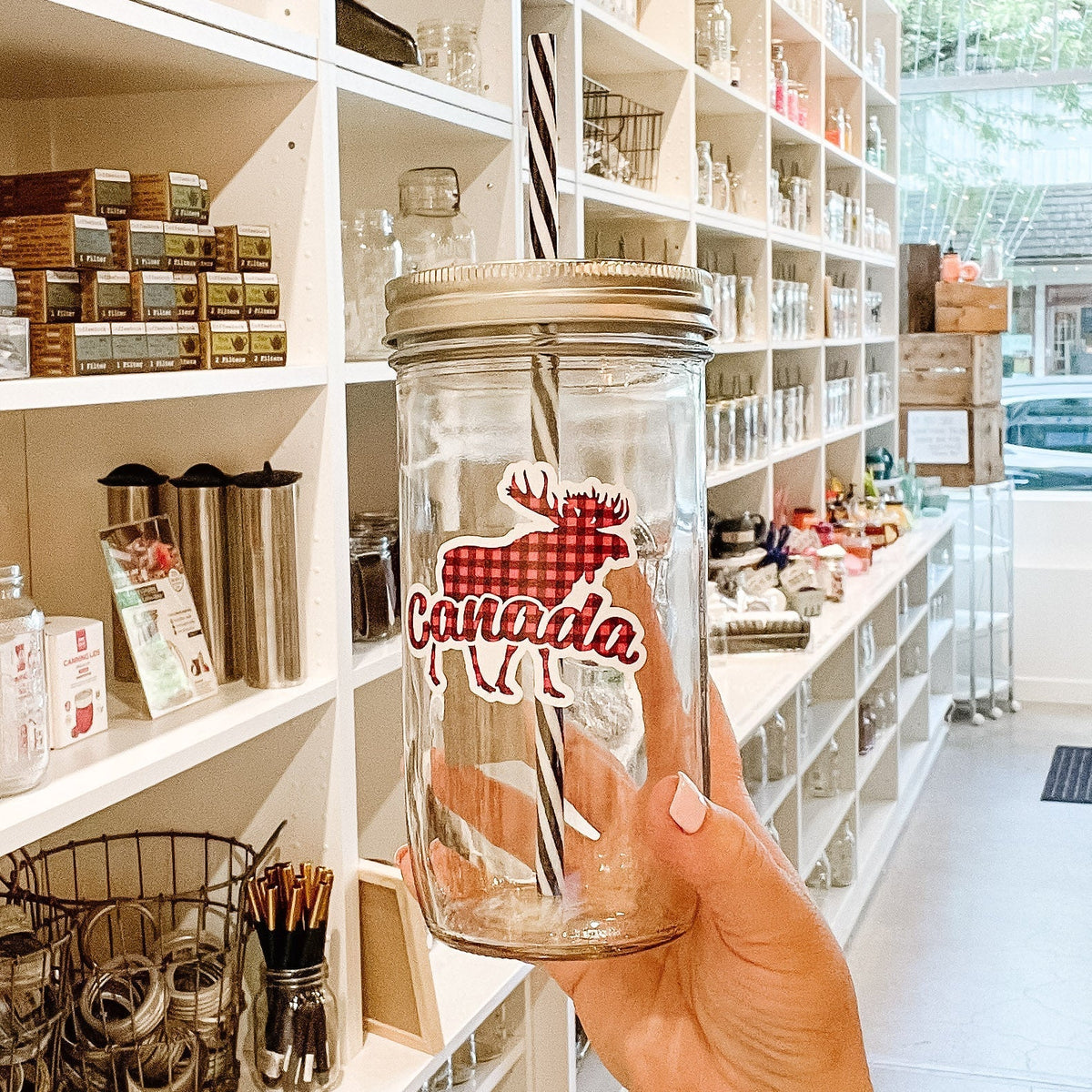 Close up of a hand holding a mason jar tumbler that says "Canada" with a silhouette of a moose behind the word. Everything in a red and black plaid pattern.
