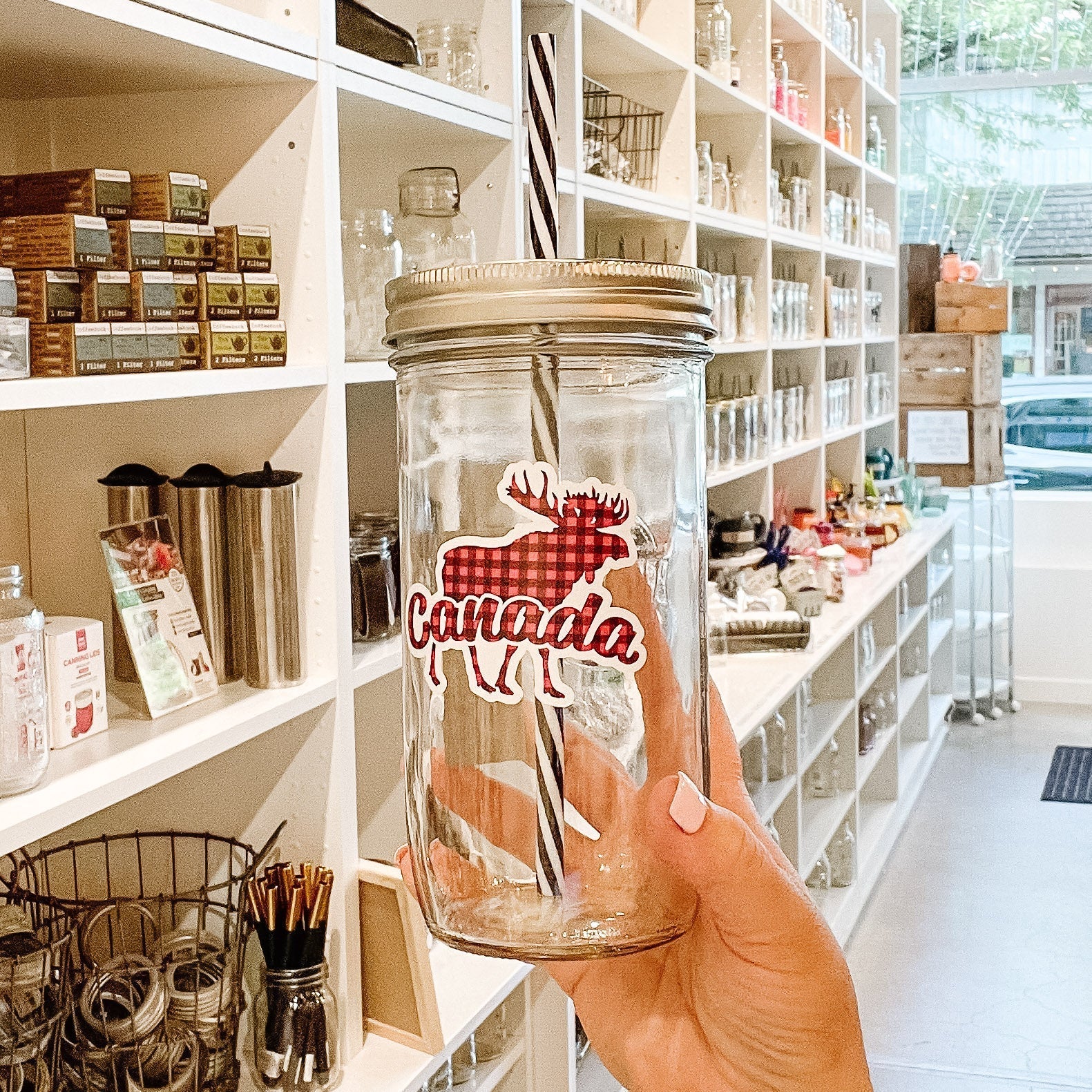 Hand holding a mason jar tumbler that says "Canada" with a silhouette of a moose behind the word. Everything in a red and black plaid pattern.