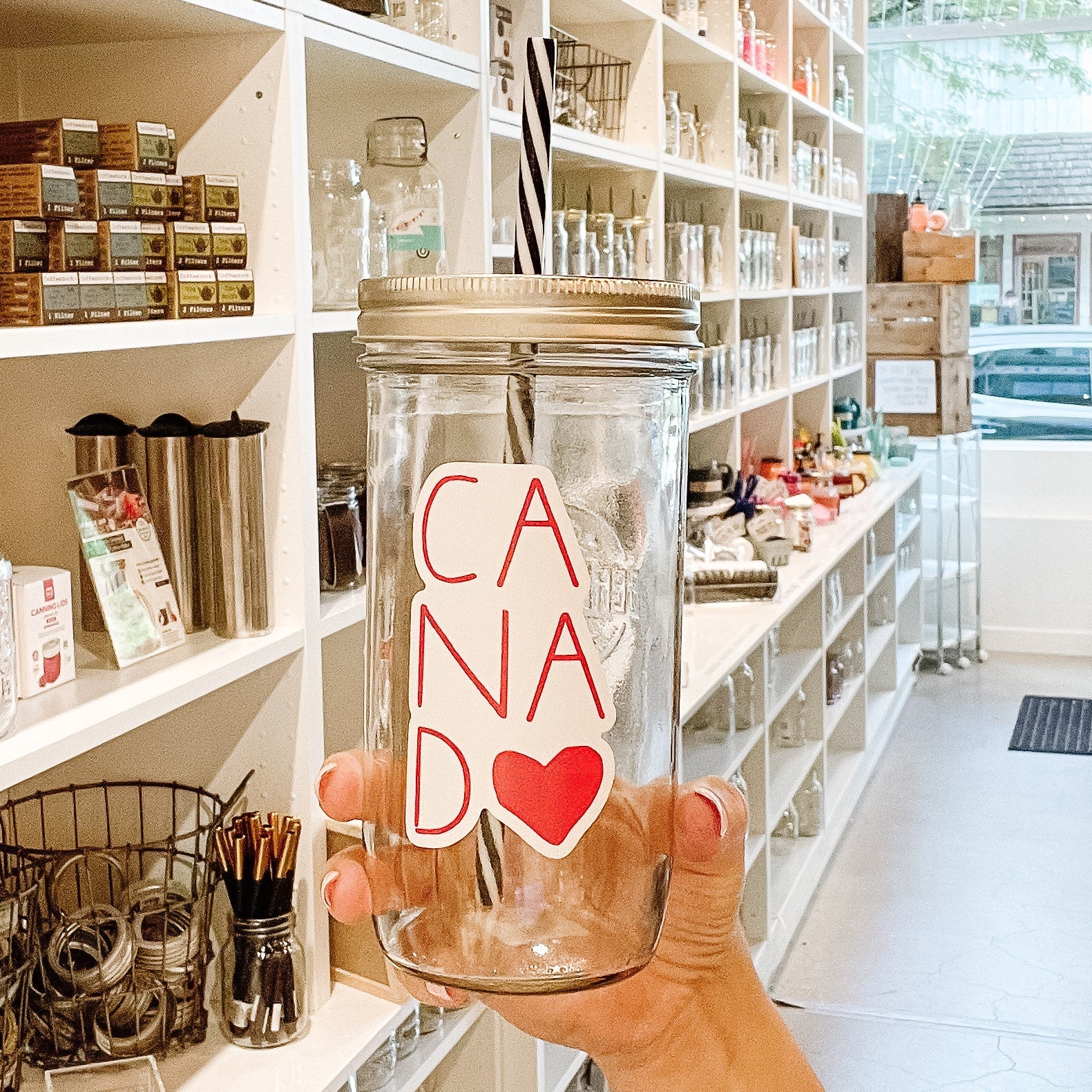 Hand holding a mason jar tumbler that says “canada” with the last A replaced by a heart. All red lettering.