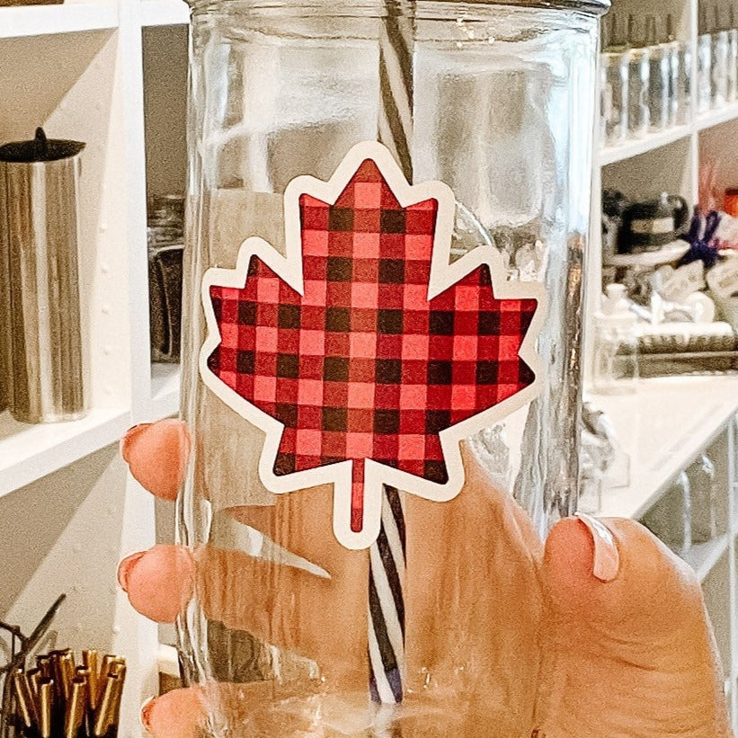 Close up of a hand holding a mason jar tumbler that has a maple leaf on it in a black and red plaid pattern.