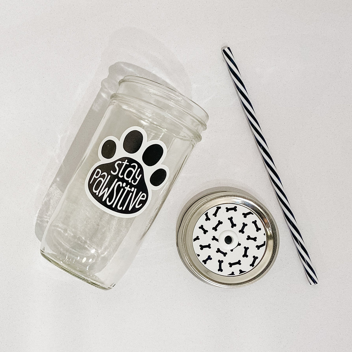 Flat lay of a mason jar tumbler that says "stay pawsitive”, a white straw lid with black dog bones repeating on it, and a black and white striped straw