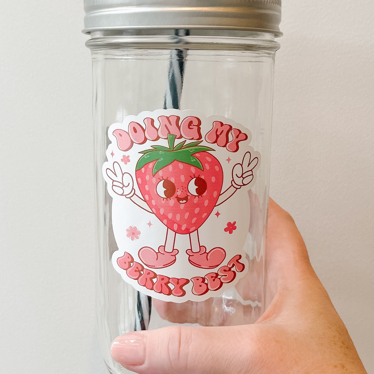 Close up of a hand holding a mason jar tumbler that says “doing my berry best” with a strawberry character holding up peace signs, the lettering is in different shades of pink.