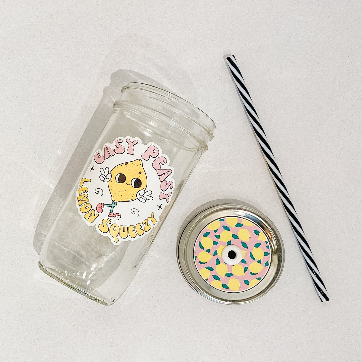 Flat lay of a mason jar tumbler that says “easy peasy lemon squeezy”, a pink straw lid with lemons all over it, and a black and white straw.