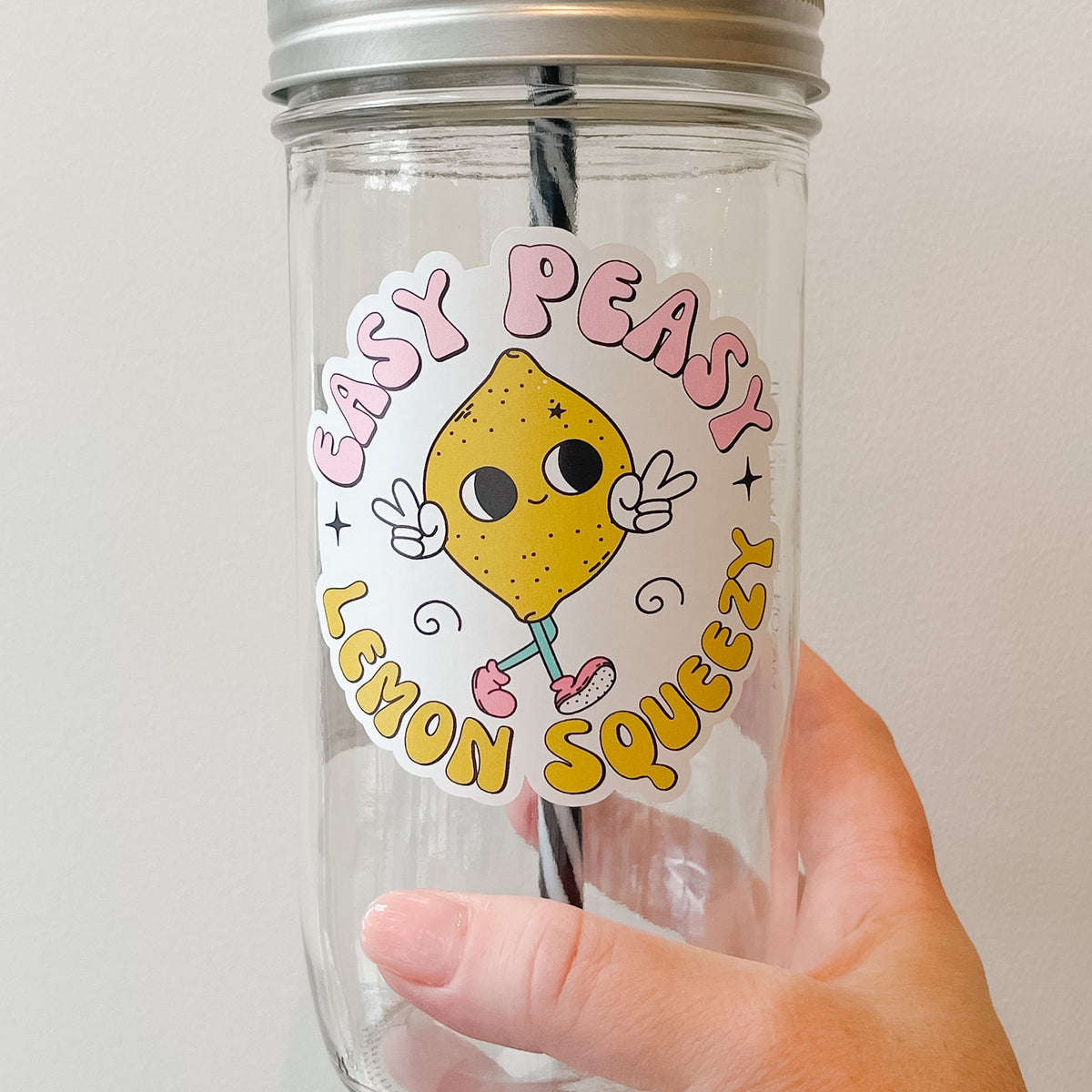 Close up of a hand holding a mason jar tumbler that says “Easy peasy lemon squeezy” with a lemon character giving 2 peace signs.