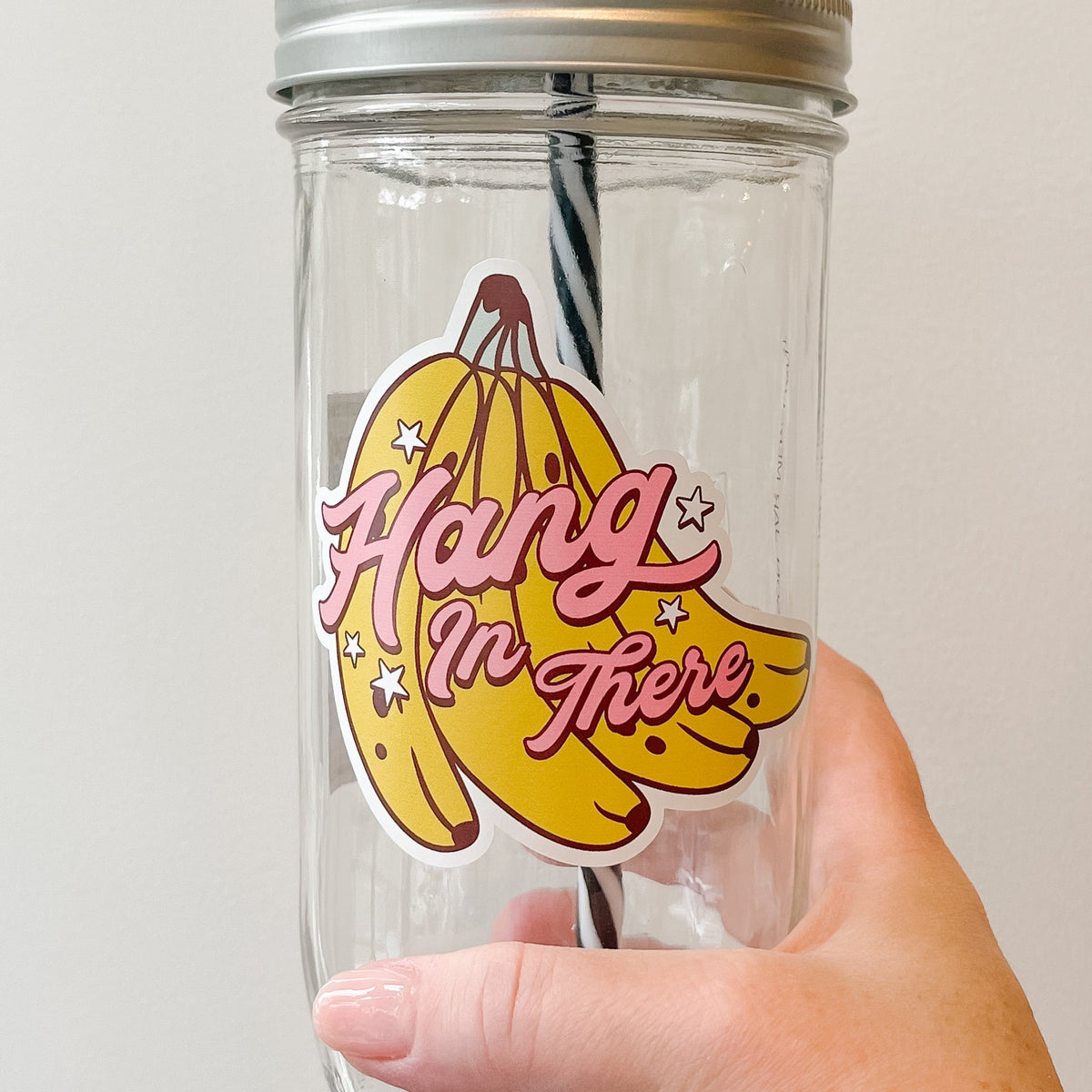 Close up of a hand holding a mason jar tumbler that says “hang in there” in pink lettering over a bunch of bananas.