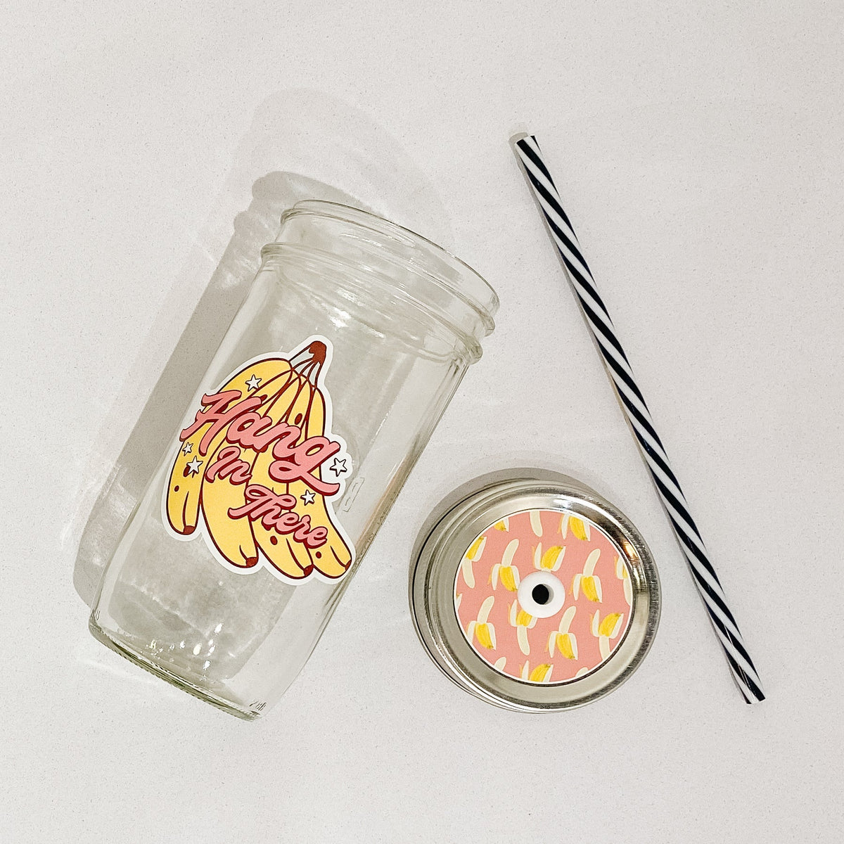 Flat lay of a mason jar tumbler that says “hang in there”, a pink straw lid with yellow bananas half-peeled, and a black and white straw.