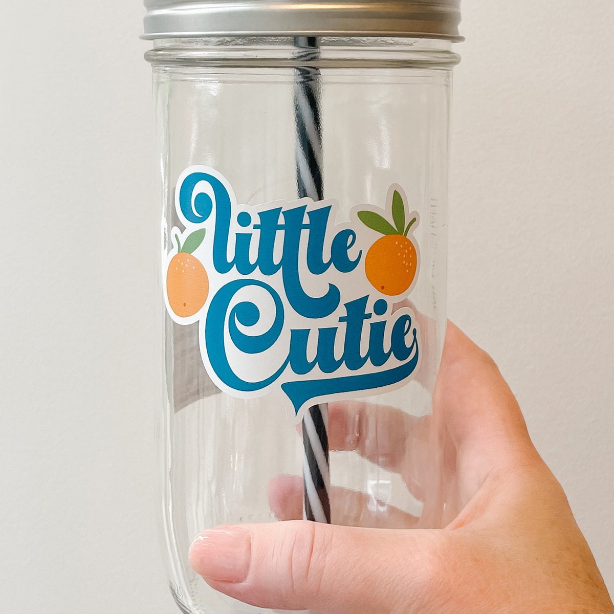 Close up of a hand holding a mason jar tumbler that says “little cutie” with two oranges on either side of the words.