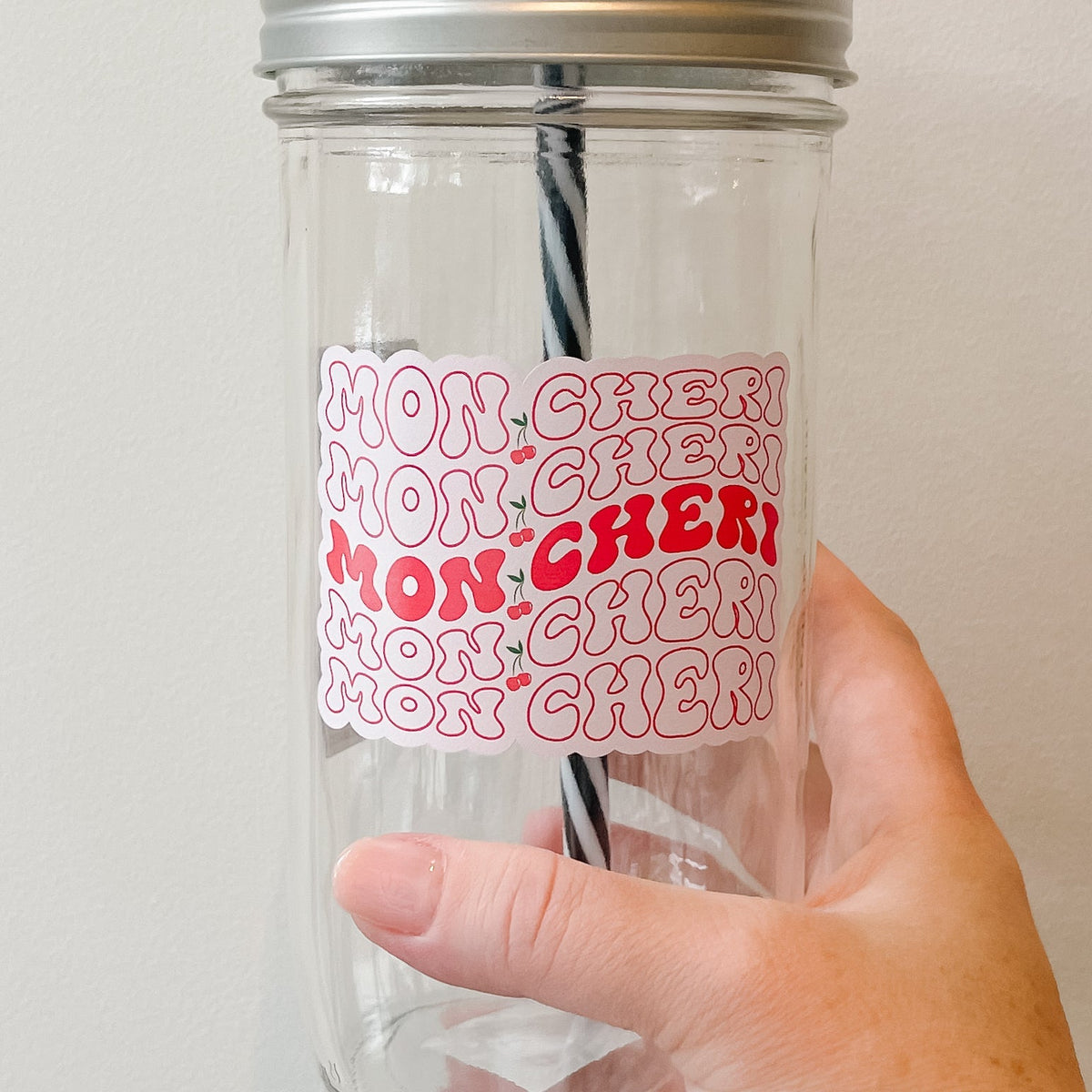 Close up of a hand holding a mason jar tumbler that says “mon cheri” with a cherry in between the words. This repeats 5 times, the words are outlined red except for the middle one where the words are filled in red. All on a pink background.