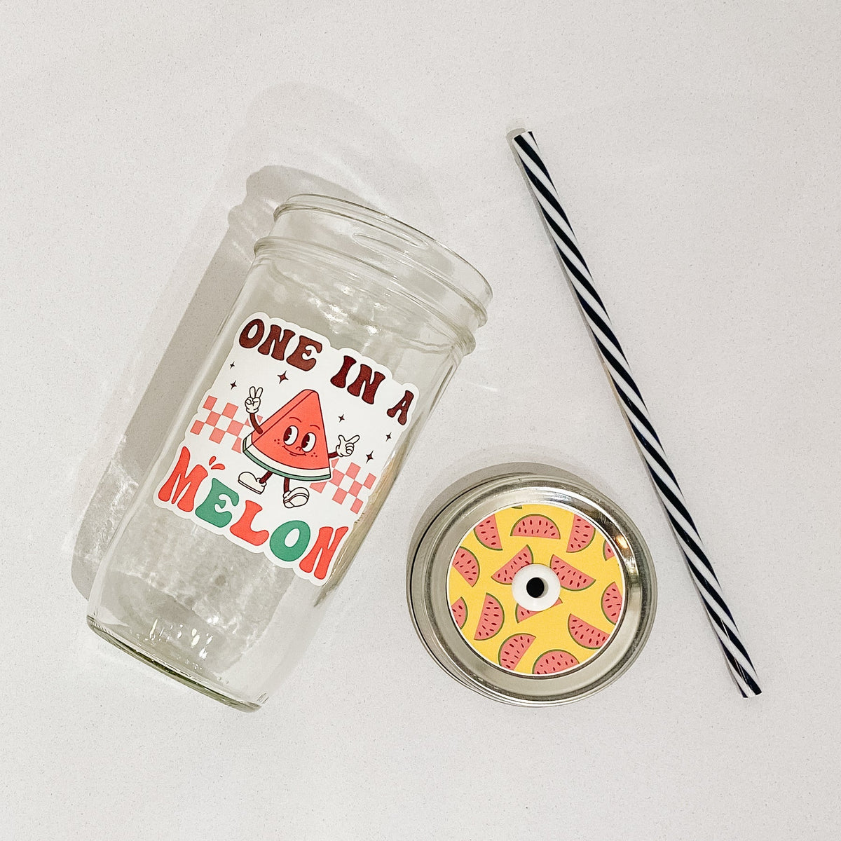 Flat lay of a mason jar tumbler that says “One in a melon”, a straw lid with pink watermelon slices on a yellow background, and a black and white striped straw.
