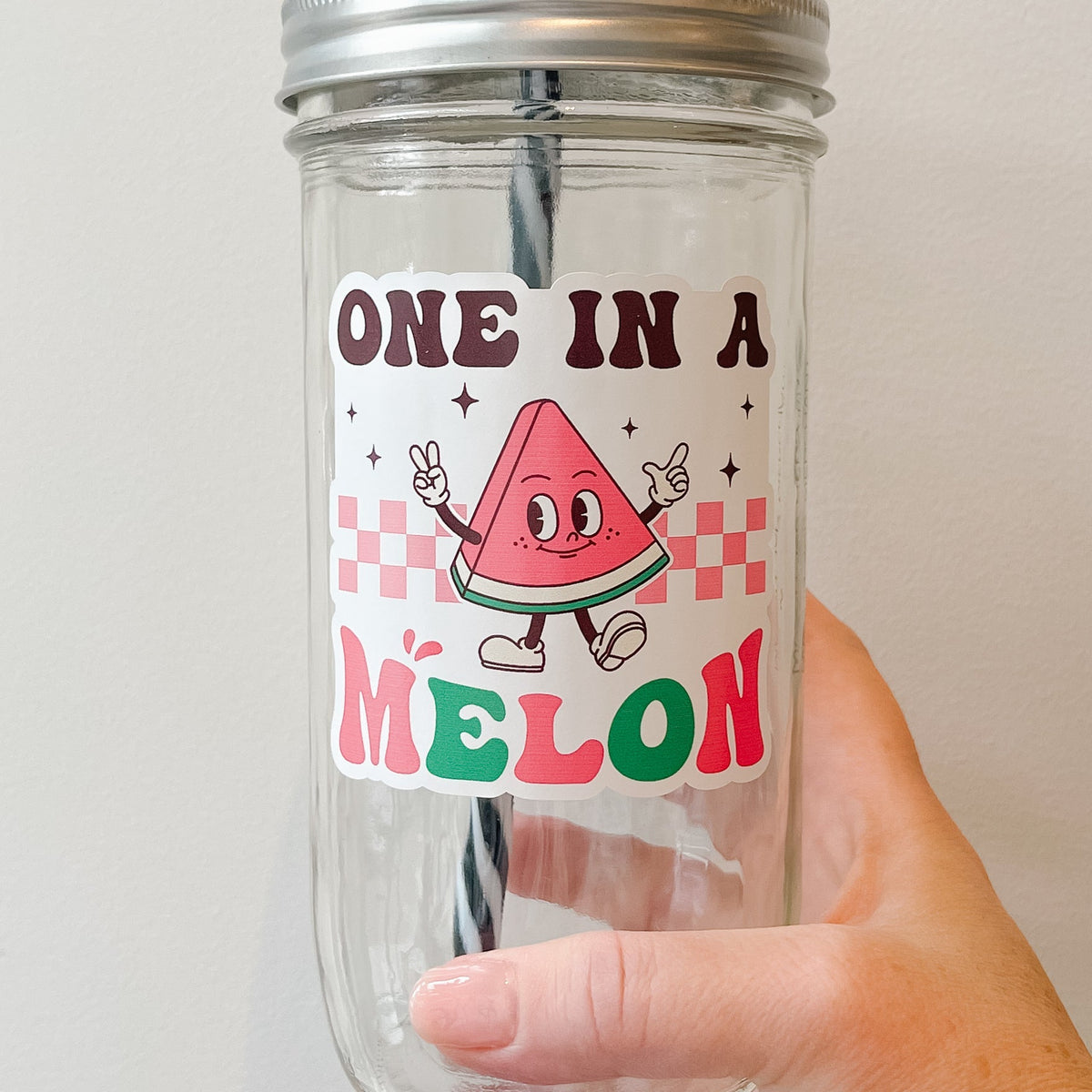 Close up of a hand holding a mason jar tumbler that says “one in a melon” with a slice of watermelon character on a white background.