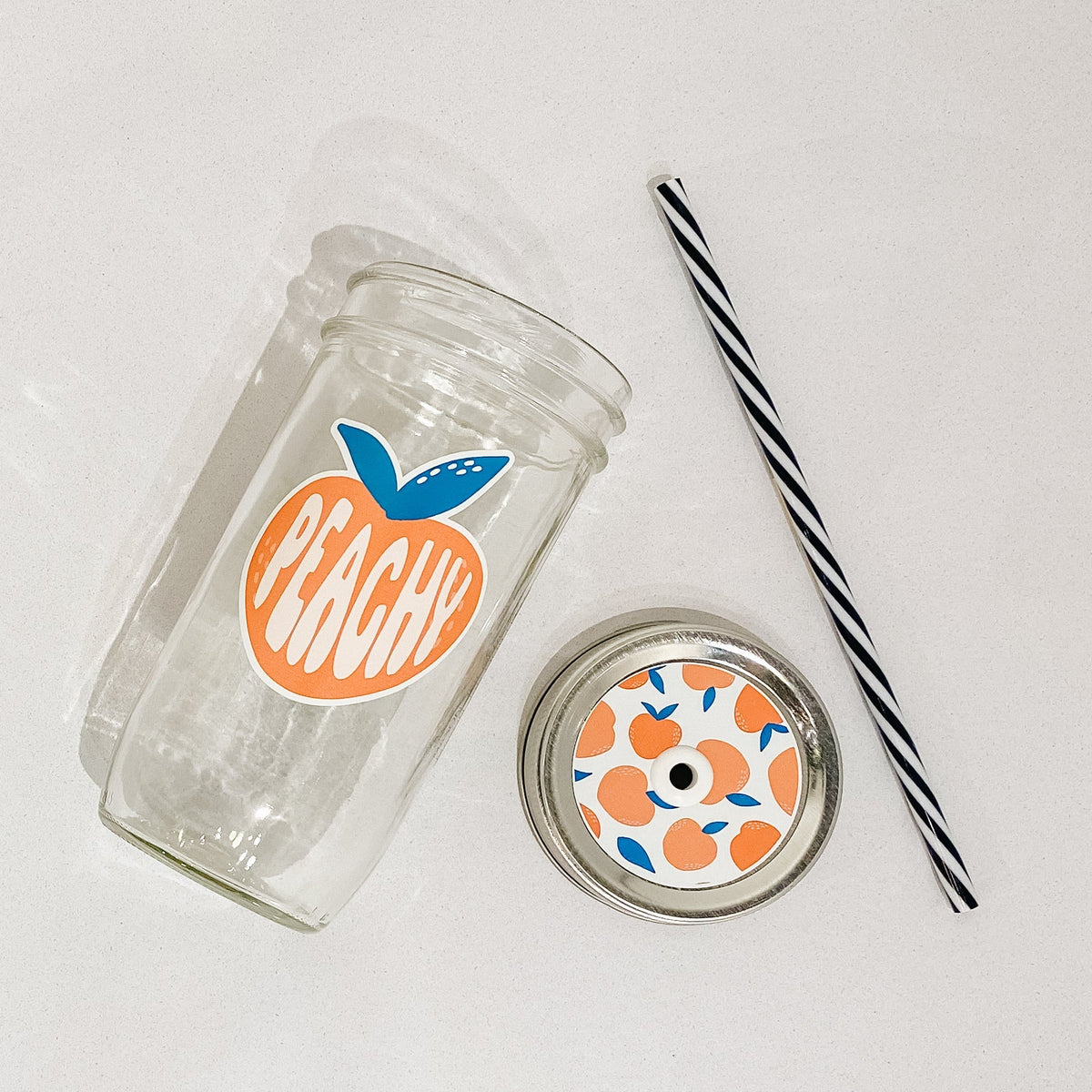 Flat lay of a mason jar tumbler that says “peachy” within a peach, a straw lid that has the same peach over it multiple times, and a black and white striped straw.
