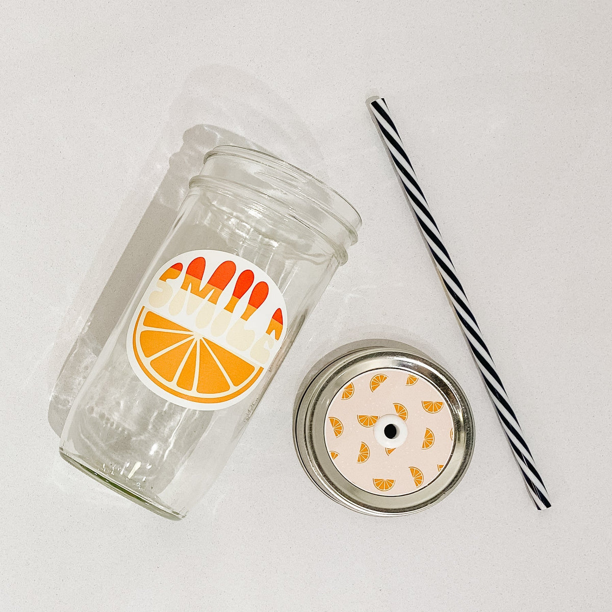 Flat lay of a mason jar tumbler that says “smile”, and straw lid that has a light peach background and orange slices all over it, and a black and white striped straw.