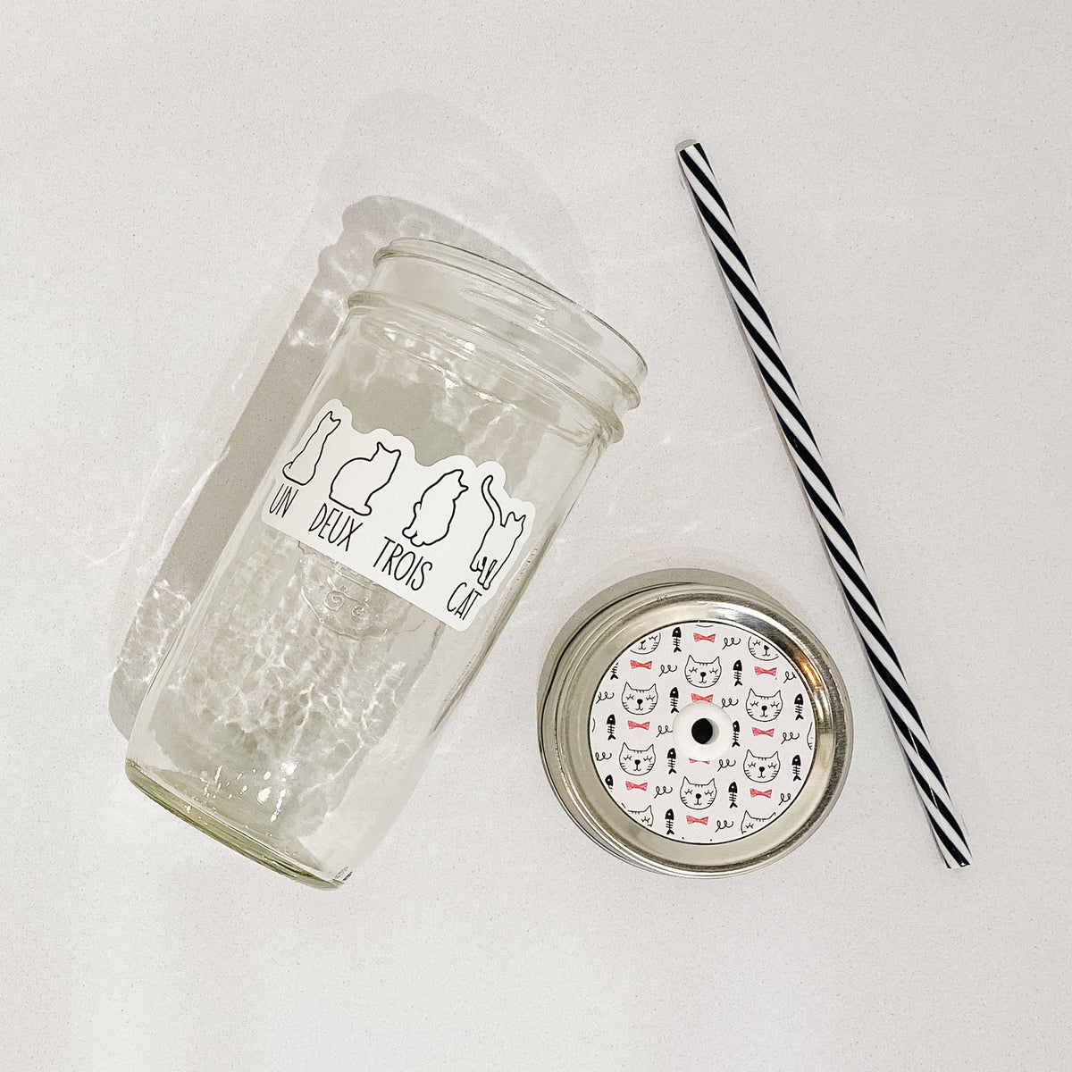 Flat lay of a mason jar tumbler that says “un deus trios cat”, a straw lid that has cat faces and fish skeletons, and a black and white striped straw