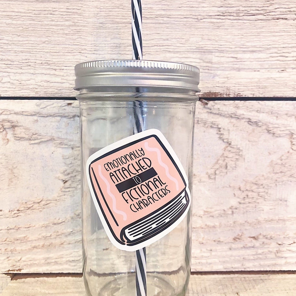 Mason jar tumbler that has an image of a pink book and the cover reads “emotionally attached to fictional characters”