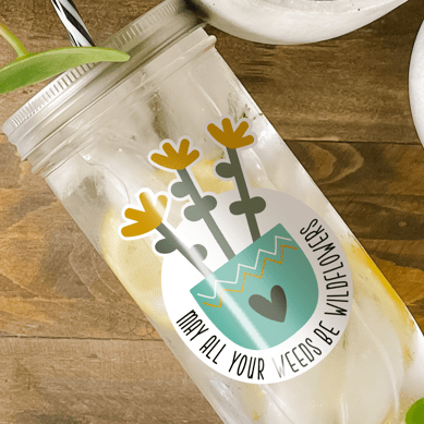 Tumbler with water inside and a sticker of wildflowers in an aqua colored pot with a heart design printed on it. There is also a print that reads "May All Your Weeds Be Wildflowers." Tumbler is photographed as a flat lay on a wooden table with some potted plants surrounding it.