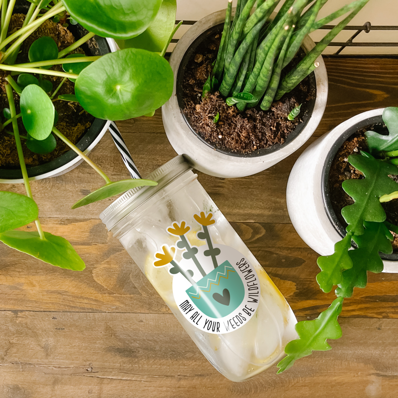 Tumbler with water inside and a sticker of wildflowers in an aqua colored pot with a heart design printed on it. There is also a print that reads &quot;May All Your Weeds Be Wildflowers.&quot; Tumbler is photographed as a flat lay on a wooden table with some potted plants surrounding it.