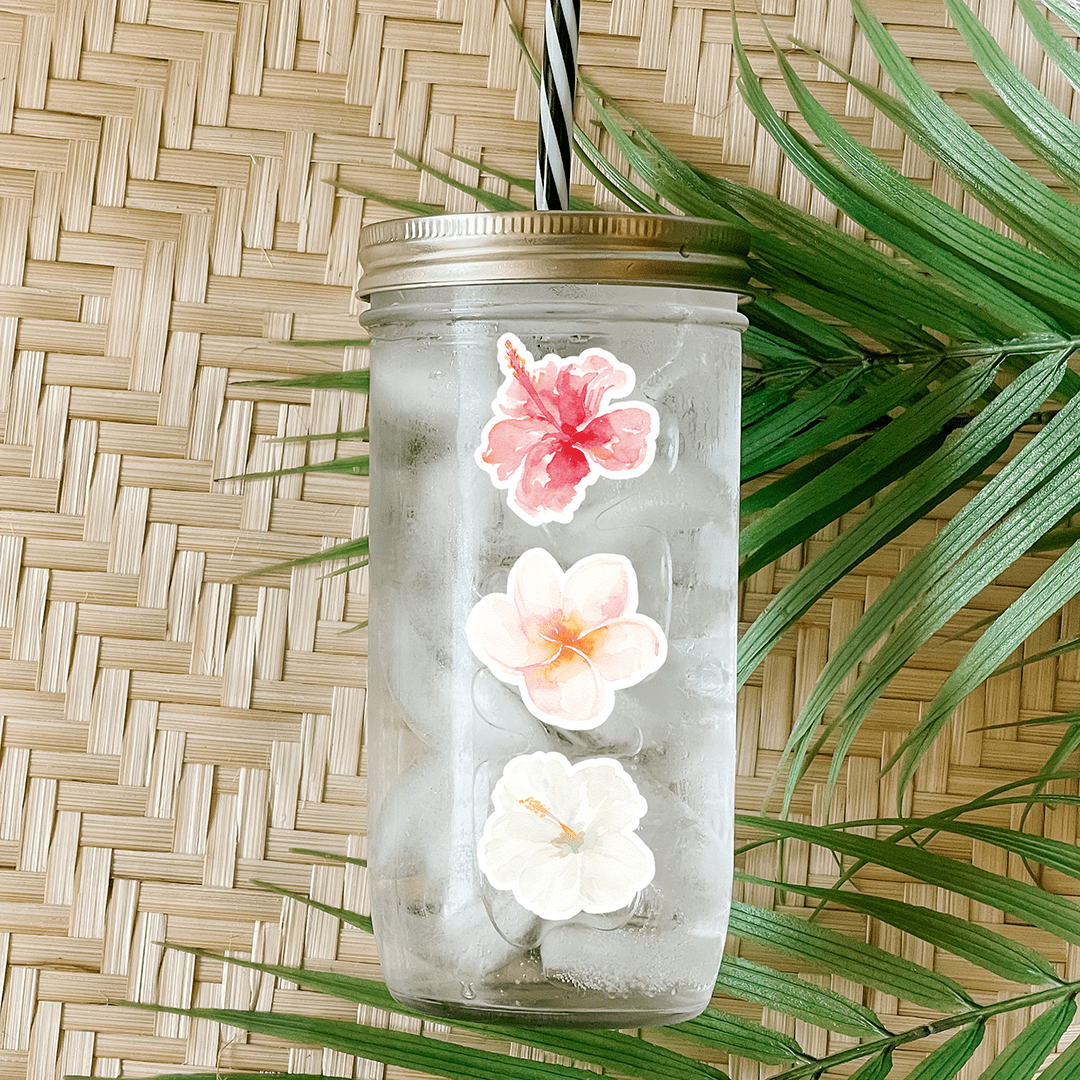 Tumbler with water and a sticker that has a pink, peach, and white hibiscus flowers. Photographed as a flat lay in a winnowing tray with palm leaves.