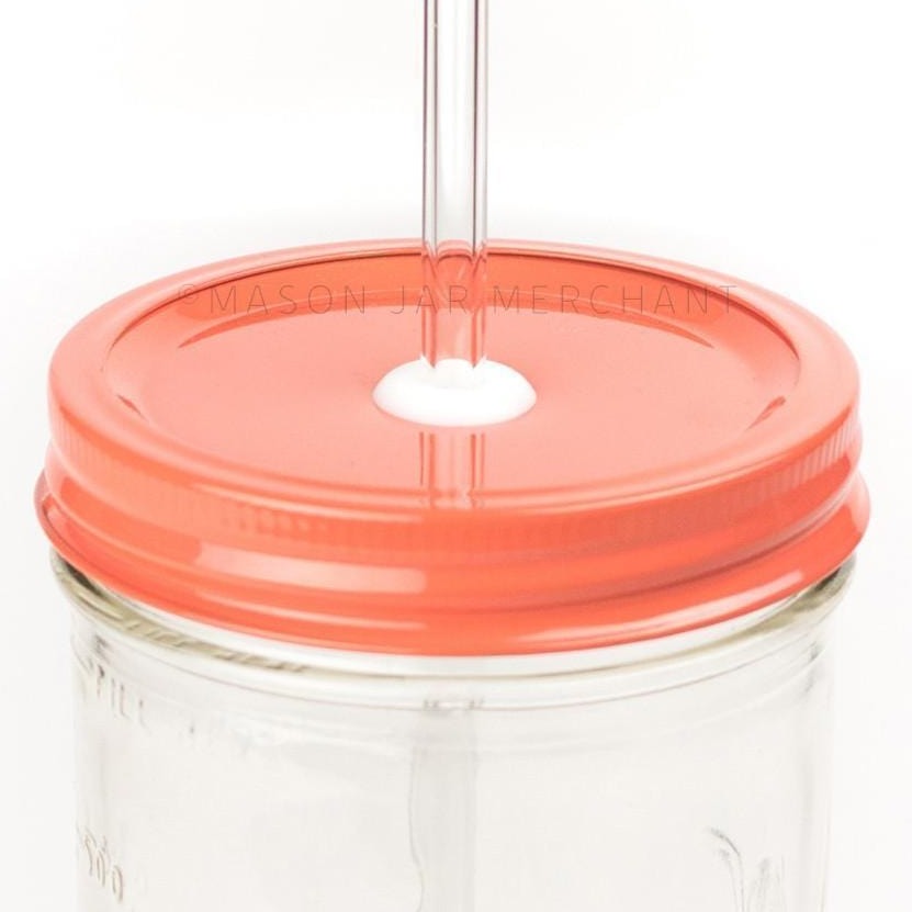 Close-up of a 24 oz mason jar with a coral coloured custom painted lid with a white silicone grommet and a glass straw, against a white background