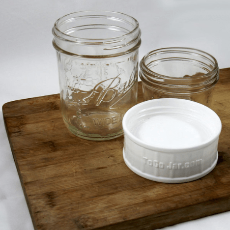 two glass reusable mason jars as well as a white to go spill proof lid sit on a cutting board on a white background