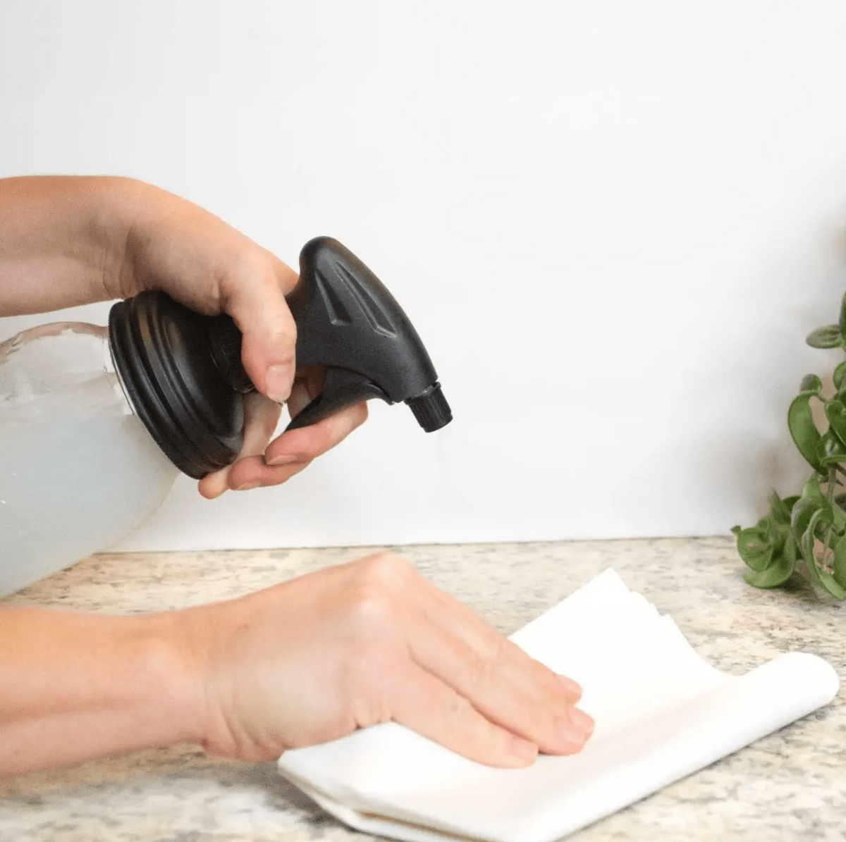Woman using a mason jar with black spray bottle lid to spray and clean a table with a tissue on the other hand.
