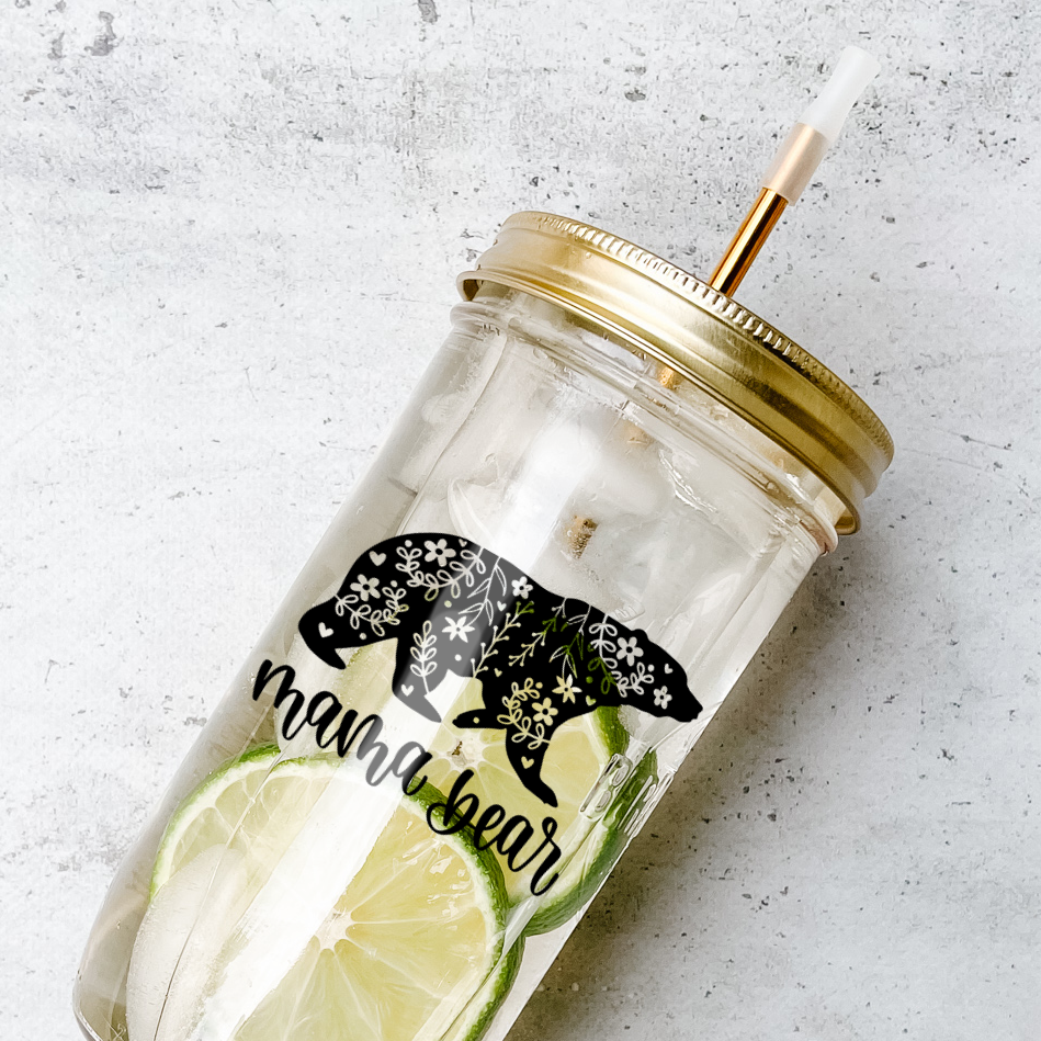 Mason jar tumbler filled with a citrus water drink and has a gold lid and straw and a calligraphy print that reads "mama bear" with white floral patterns in a black bear graphic. Photographed against a marble countertop.