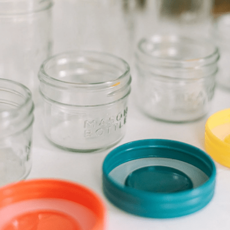 Close up of 4oz mason jar bottles and multicolored plastic lids in a white countertop.