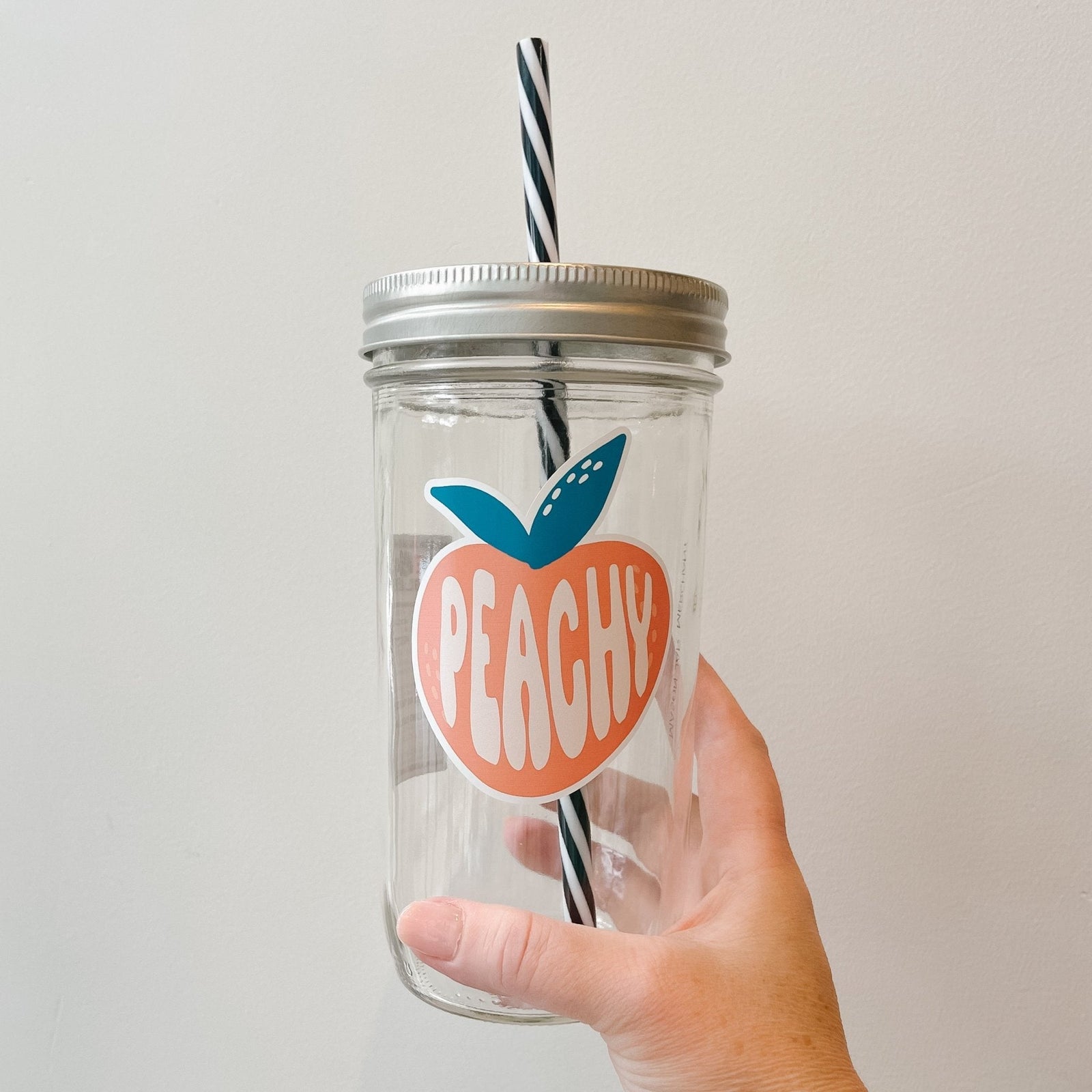 Hand holding a mason jar tumbler that has an image of a cartoon-ish peach with the word “peachy” within the fruit.