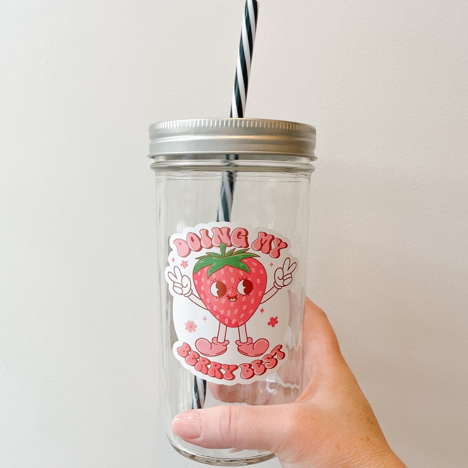 Hand holding a mason jar tumbler that says “doing my berry best” with a strawberry character holding up peace signs, the lettering is in different shades of pink.
