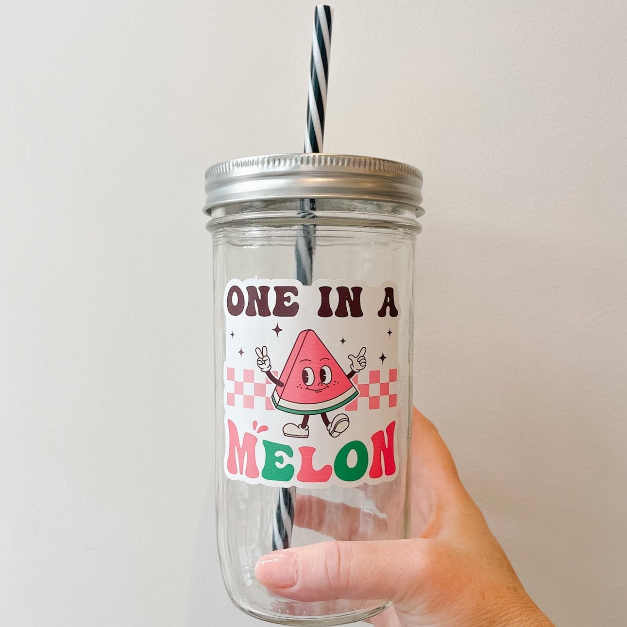 Hand holding a mason jar tumbler that says “one in a melon” with a slice of watermelon character on a white background. 