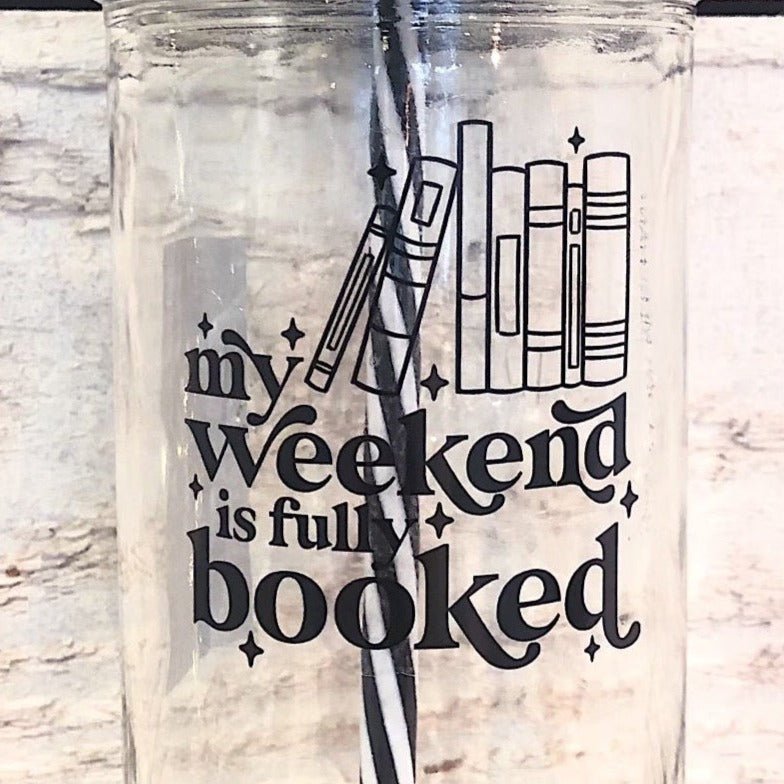 Flat lay of a mason jar tumbler that says “my weekend is fully booked”, a straw lid that is a bookshelf full of multicoloured books, and a black and white striped straw