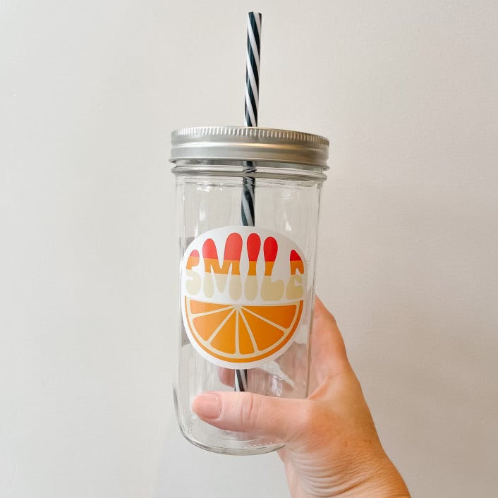 A hand holding a clear glass mason jar tumbler with a silver lid and a black-and-white striped straw. The jar features a circular graphic with the word 'SMILE' in red and orange above an orange slice illustration.