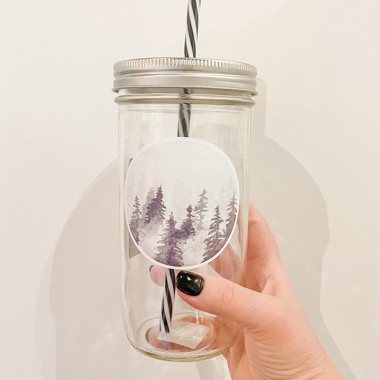 Hand holding a mason jar tumbler that has a circle decal of trees in different shades of grey in fog. 