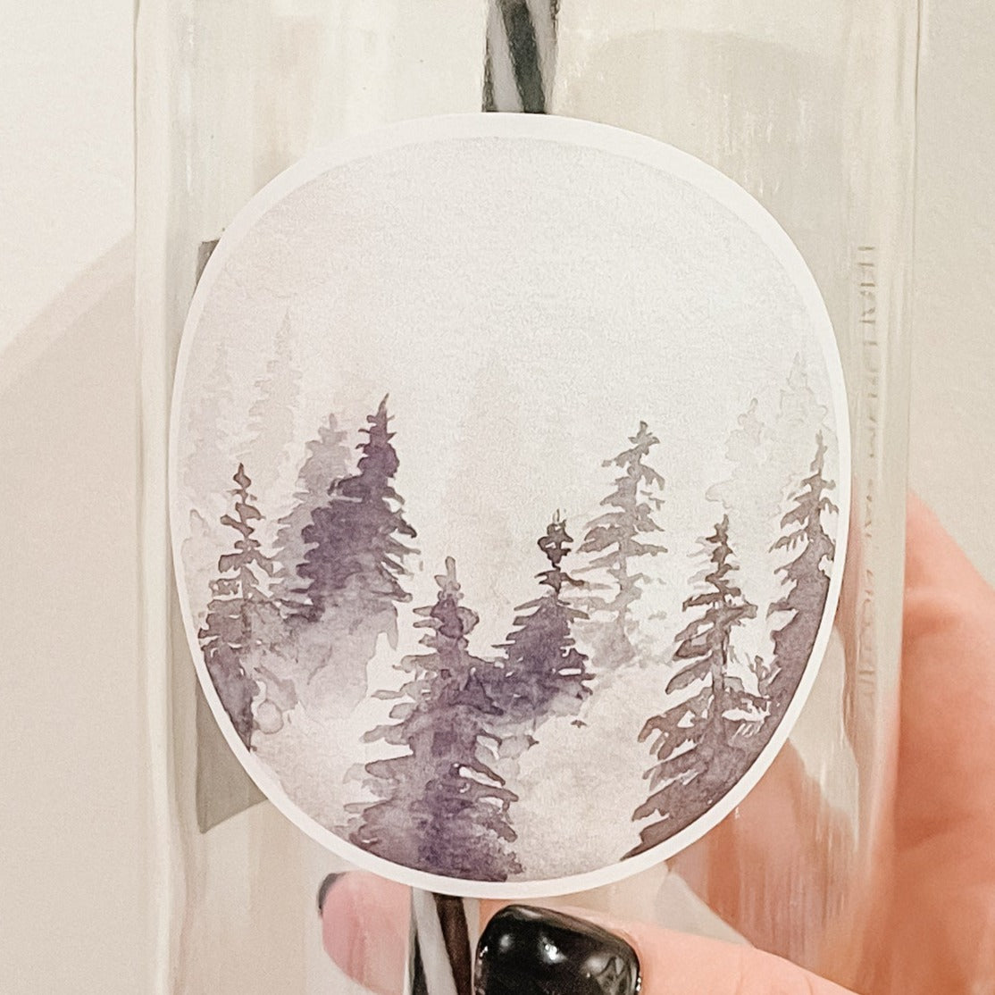 Hand holding a mason jar tumbler that has a circle decal of trees in different shades of grey in fog. 