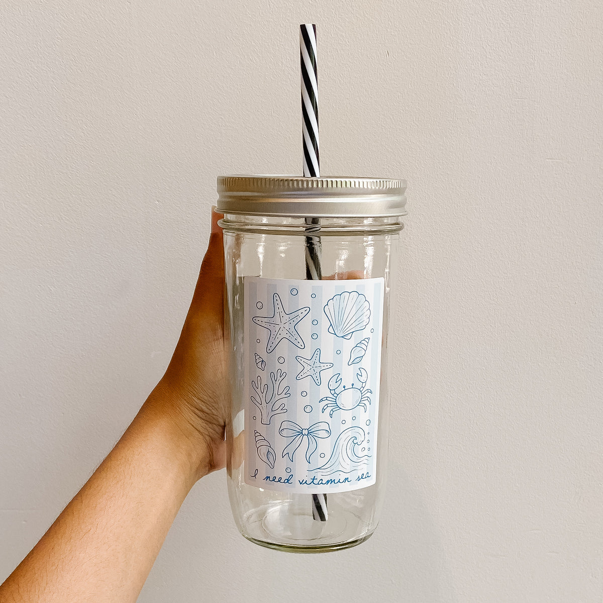 Hand holding mason jar tumbler that reads i need vitamin sea with soft blue beachy  stripe design and paired with a black and white striped straw 