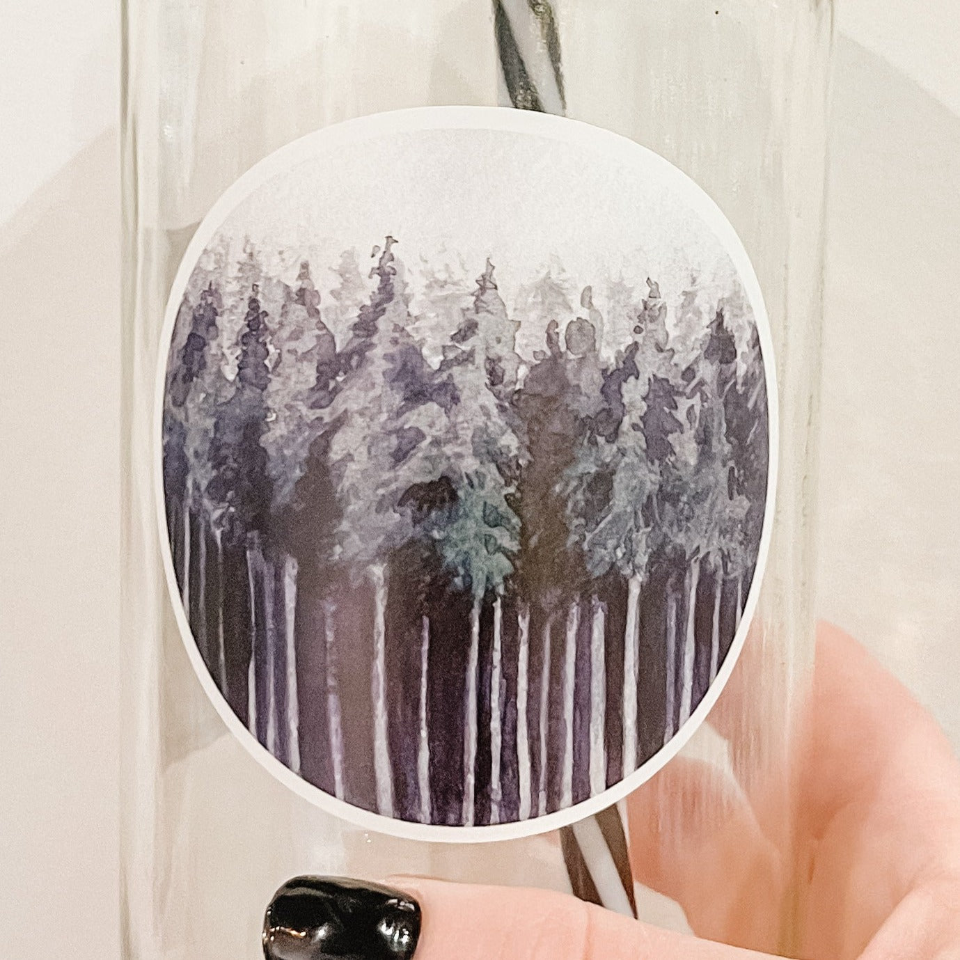 Hand holding a mason jar tumbler with a circular decal of multiple trees in shades of dark green and greys. 