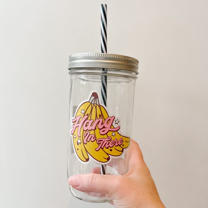 A hand holding a clear glass mason jar tumbler with a silver metal lid and a black-and-white striped straw. The jar features a graphic of yellow bananas with the pink text 'Hang in There'.