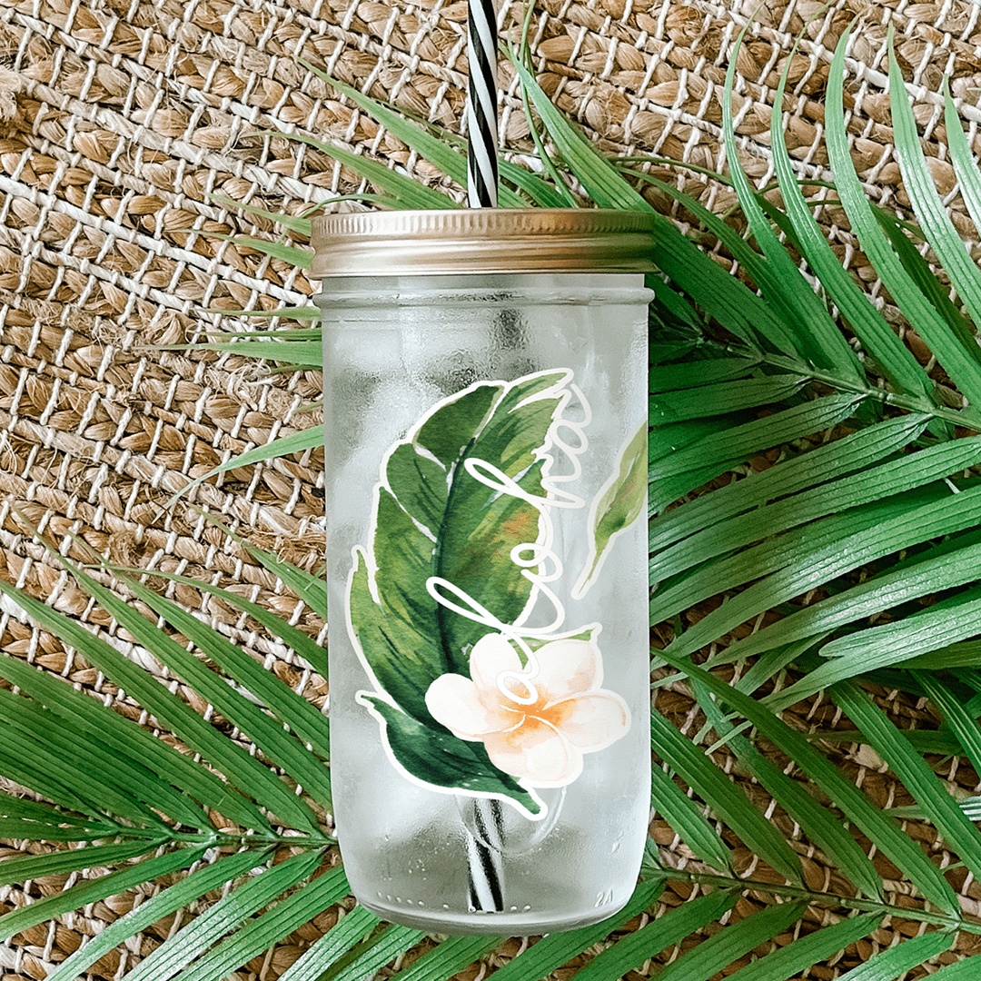 Tumbler with water and a sticker that reads "Aloha" tropical leaf and white hibiscus flower. Photographed as a flat lay in a weave mat and palm leaves.
