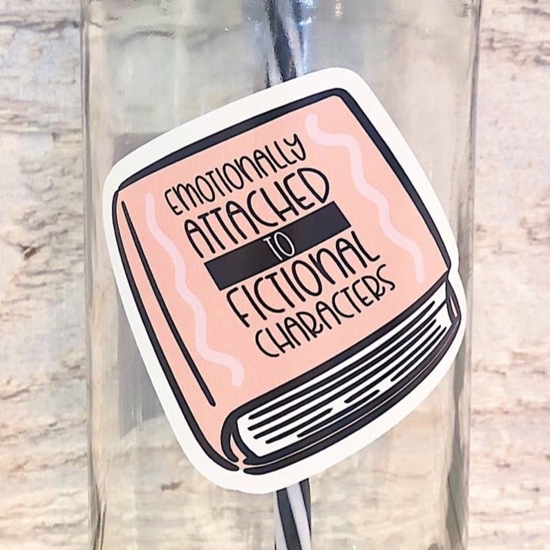 Flat lay of a mason jar tumbler that has an image of a book that says “emotionally attached to fictional character”, a pink straw lid with a repeating pattern of an opened book, and a black and white striped straw. 