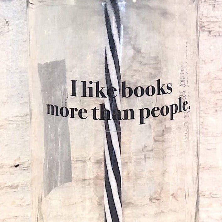 Flat lay of a mason jar tumbler that says "I like books more than people”, a straw lid this a bookshelf scene on it, and a black and white striped straw. 