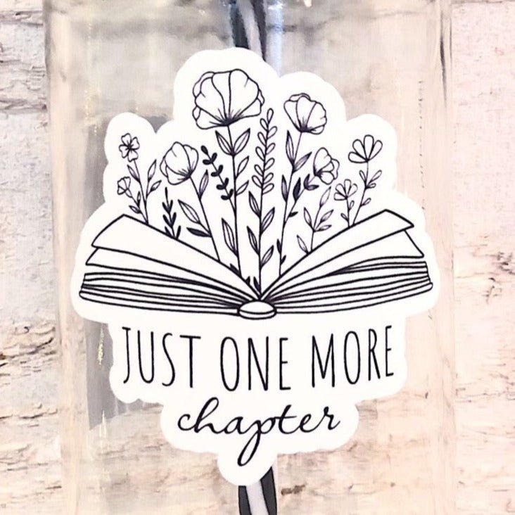 Flat lay of a mason jar tumbler that says “just one more chapter” with flowers growing out of an open book, a straw lid that has a repeating pattern of a green book, and a black and white striped straw.