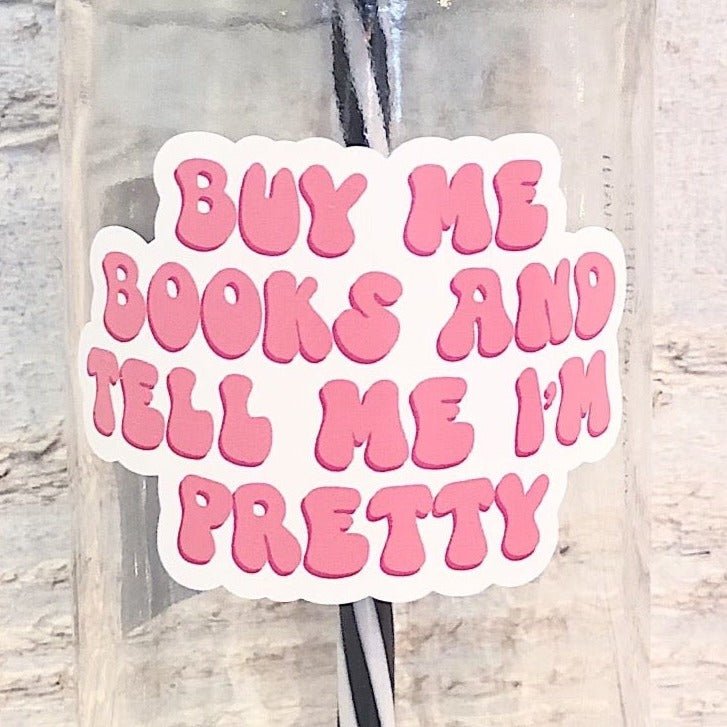 Flat lay of a mason jar tumbler that says “buy me books and tell me I’m pretty”, a pink straw lid with a repeating pattern of an opened book, and a black and white striped straw. 
