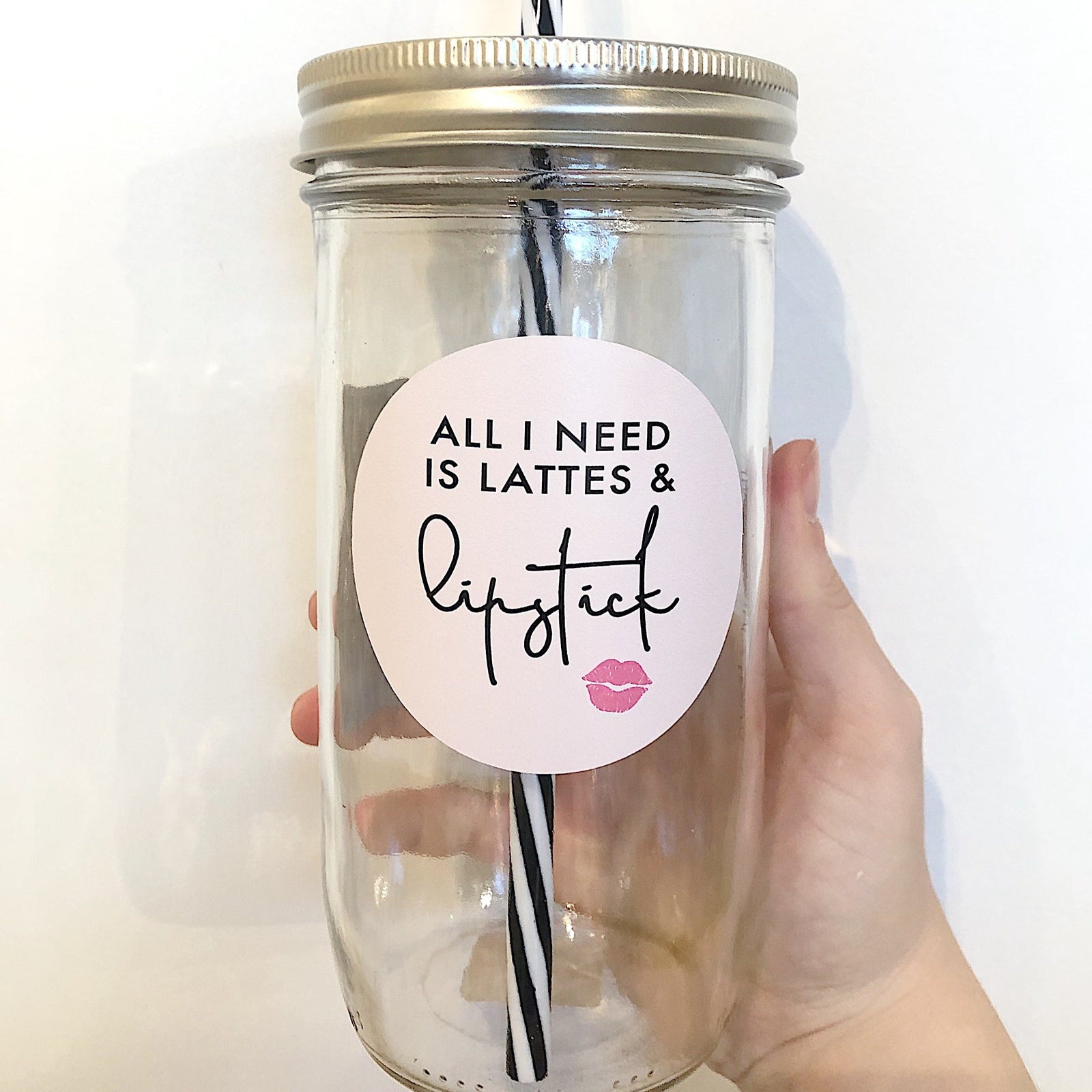 Hand holding a mason jar tumbler that says “All I need is lattes & lipstick” with a small lip kiss under. The words are black on a circular decal that is light pink. 