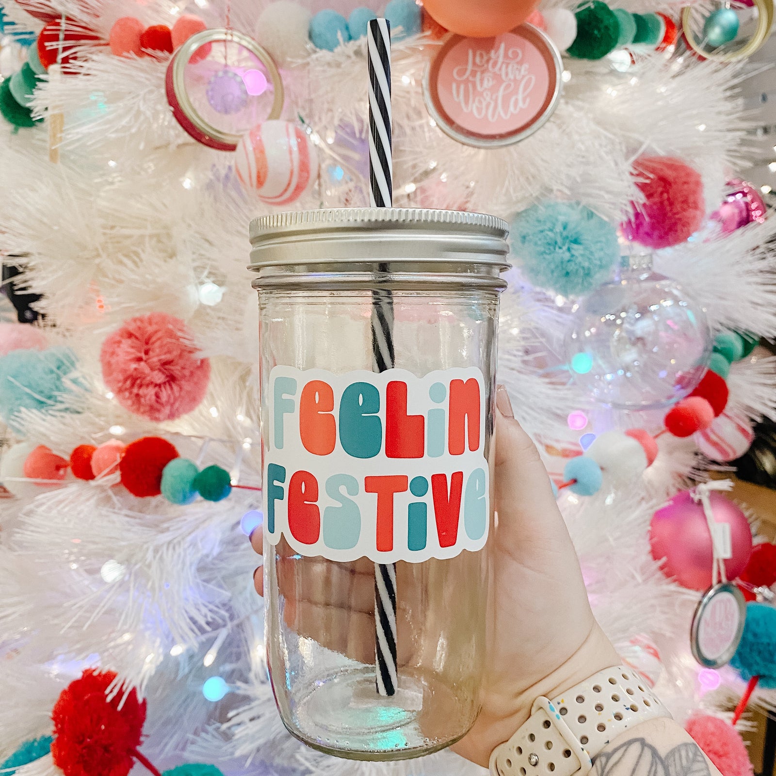 Hand holding  mason jar Tumbler that reads feelin’ festive with blues, reds and teals for the font colors. Black and white striped plastic straw inside with Christmas tree in background.