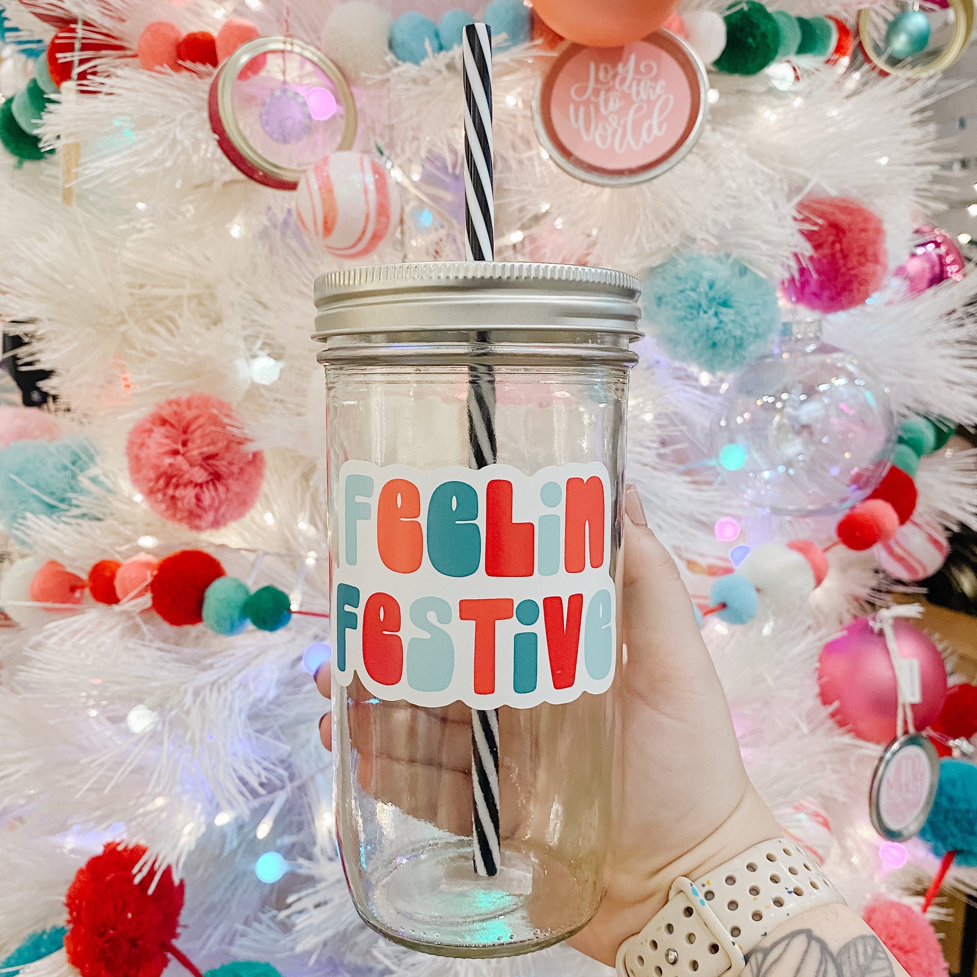 Hand holding  mason jar Tumbler that reads feelin’ festive with blues, reds and teals for the font colors. Black and white striped plastic straw inside with Christmas tree in background.