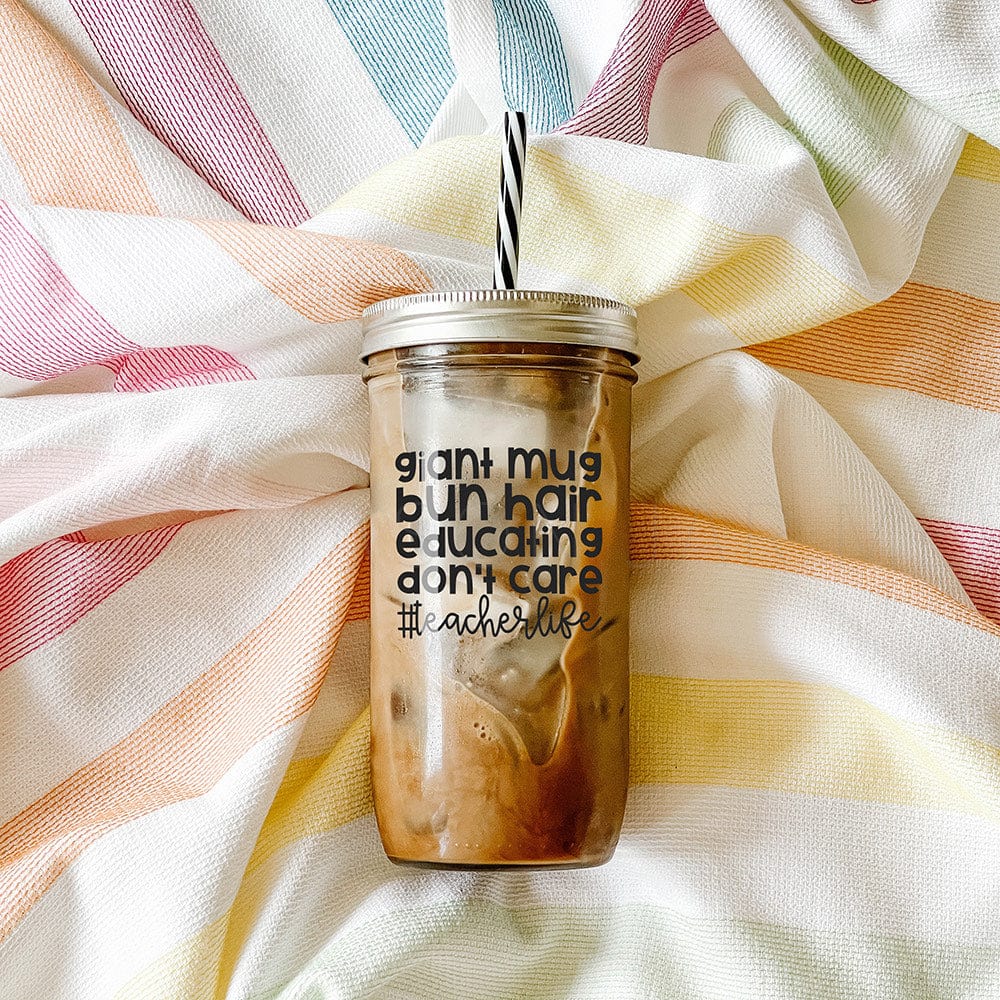 A #TeacherLife Mason Jar Tumbler with text that says "giant mug bun hair educating don't care". It has an iced coffee inside