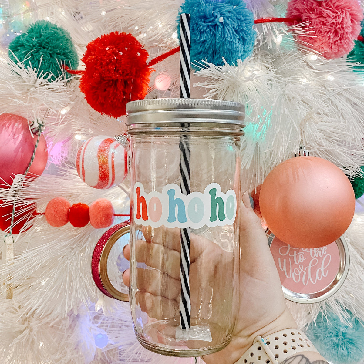 Hand holding mason jar Tumbler that reads Hohoho with blues, reds, pinks and teals for the font colors. Black and white striped plastic straw inside with Christmas tree in background.