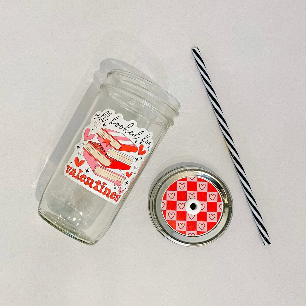 hand holding a mason jar tumbler that says "all booked for valentines" with a stack of books with red and pink covers,  and red and pink hearts around