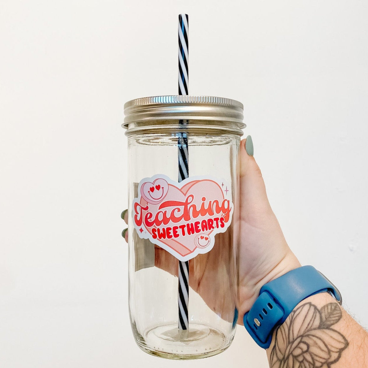 hand holding a mason jar tumbler that says "Teaching Sweethearts" with a heart behind the words and two smiley faces with hearts for eyes