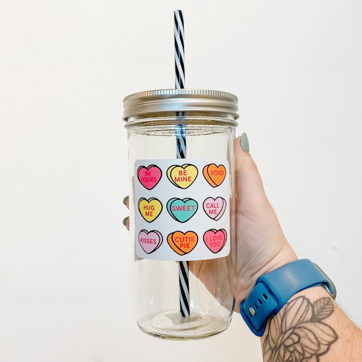 hand holding a mason jar tumbler that has 9 hearts that depict different "sweethearts" candy hearts with different valentines sayings