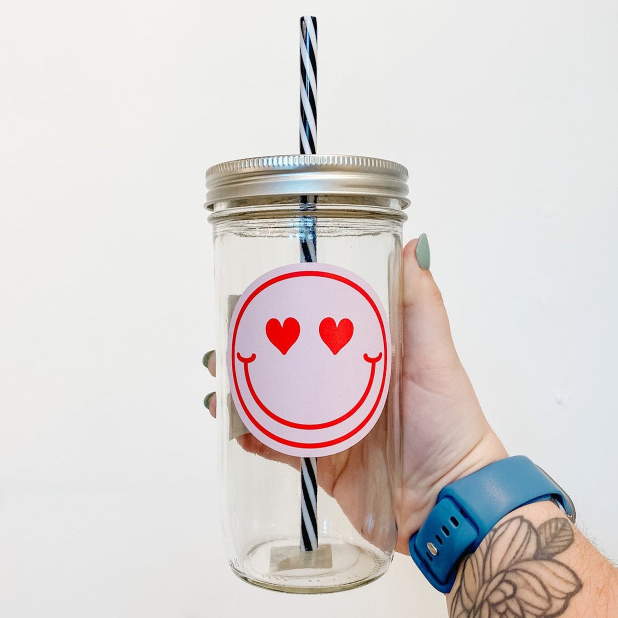 hand holding a mason jar tumbler that has a pink circular decal with a red smiley face on it with hearts for eyes